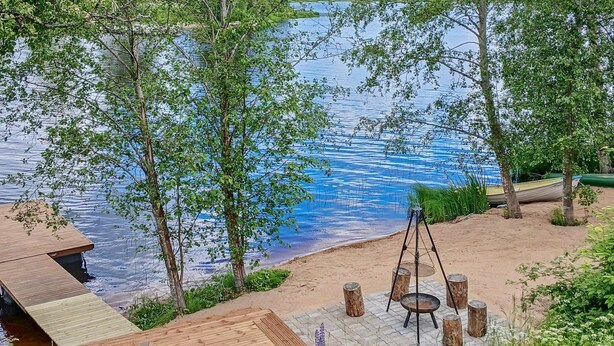A wooden deck with a metal table and chairs, overlooking a lake with trees and a hammock.