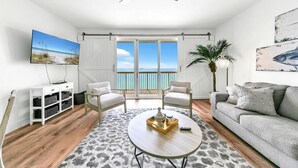A living room with a view of the ocean, a ceiling fan, a TV, a white cabinet, two chairs, a gray sofa, and a coffee table.