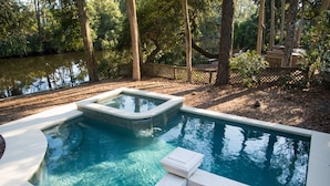 A rectangular swimming pool with a small hot tub, surrounded by a wooden deck and a fence, with trees and a body of water in the background. - Private Home with SPA and Private pool, Beautiful Lagoon views (Hilton Head Island)