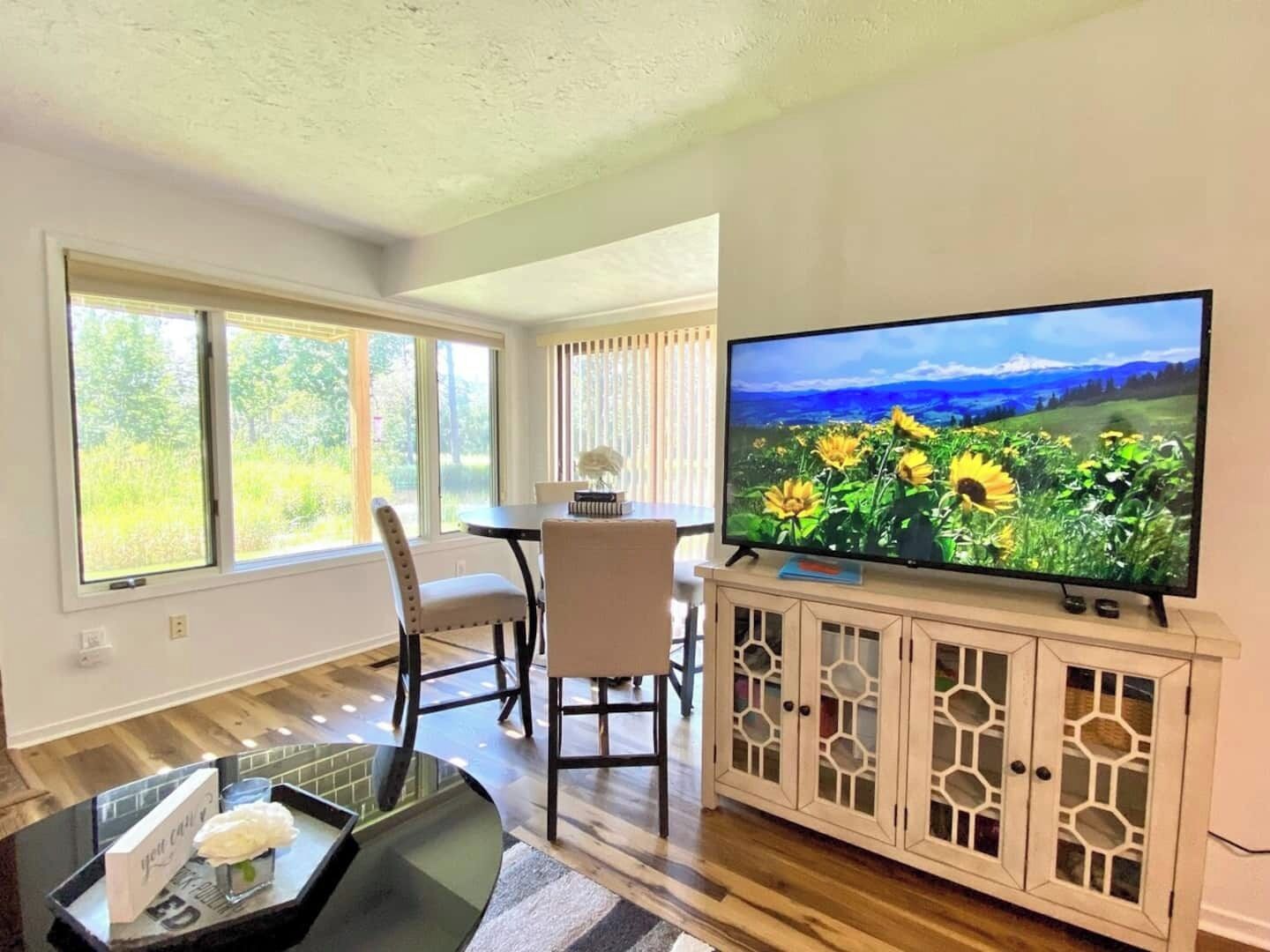 A living room with a TV displaying a sunflower field, a dining area with a table and chairs, and large windows with curtains.