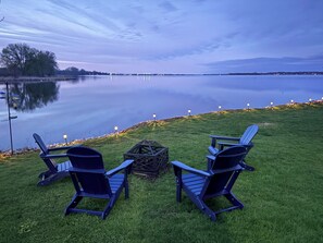 Dos sillas Adirondack mirando a un lago tranquilo con una orilla lejana y un cielo despejado.