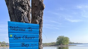 A wooden sign reading "Welcome to Sun Chaser Bay" attached to a tree by a lake with a boat and flowers in the foreground. - A Fisherman’s and birdwatchers Paradise! (Carrying Place)