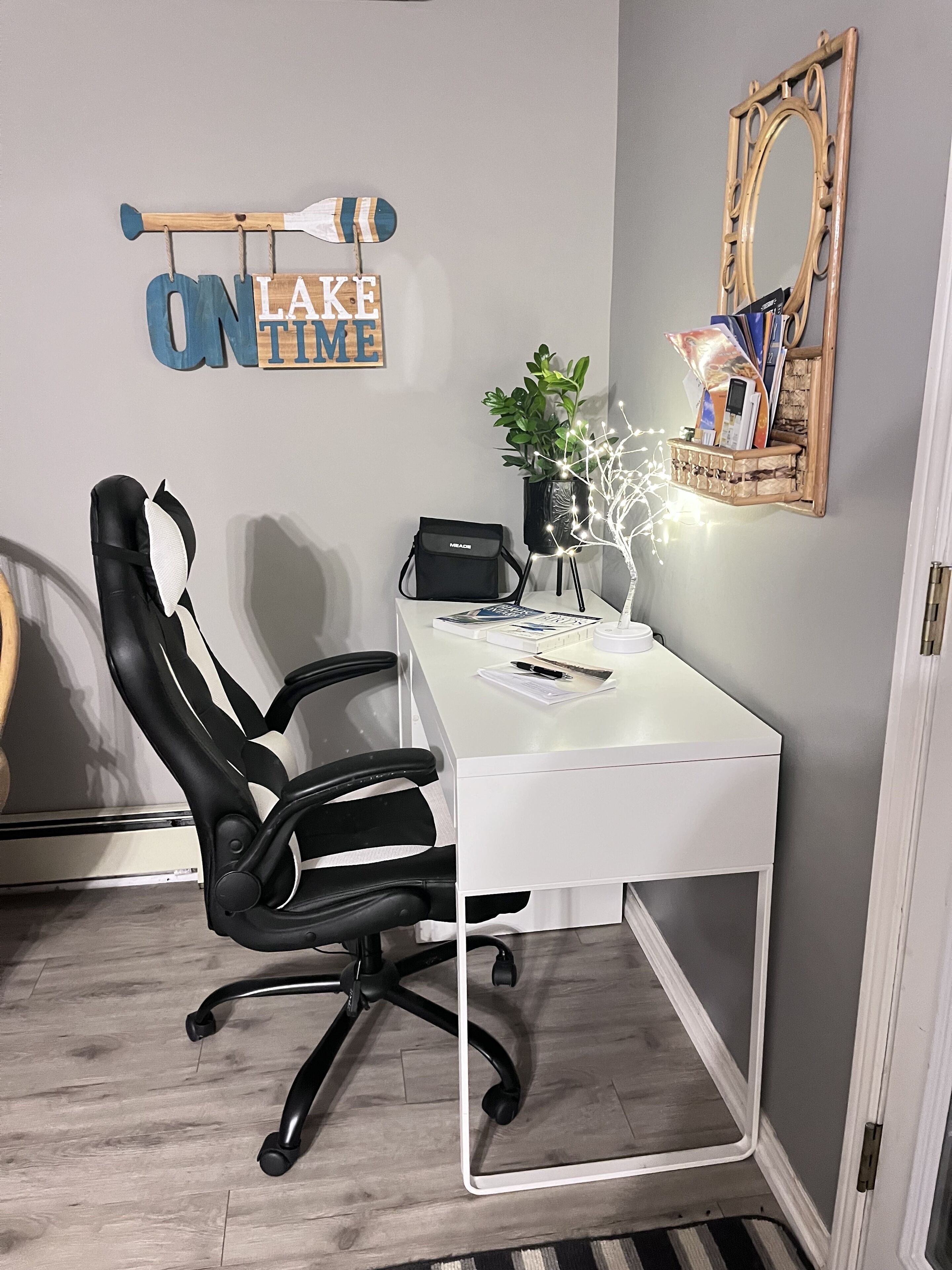 Un espace de bureau moderne avec un bureau blanc, une chaise de bureau noire, une plante en pot, une petite étagère avec des livres et un miroir.