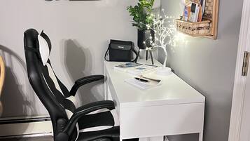 Un espace de bureau moderne avec un bureau blanc, une chaise de bureau noire, une plante en pot, une petite étagère avec des livres et un miroir.