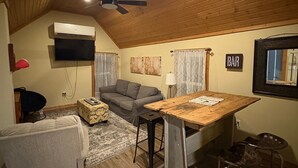 A cozy, compact living space with a wooden ceiling, a ceiling fan, a flat-screen TV, a sofa, a small dining table, and a bar area.