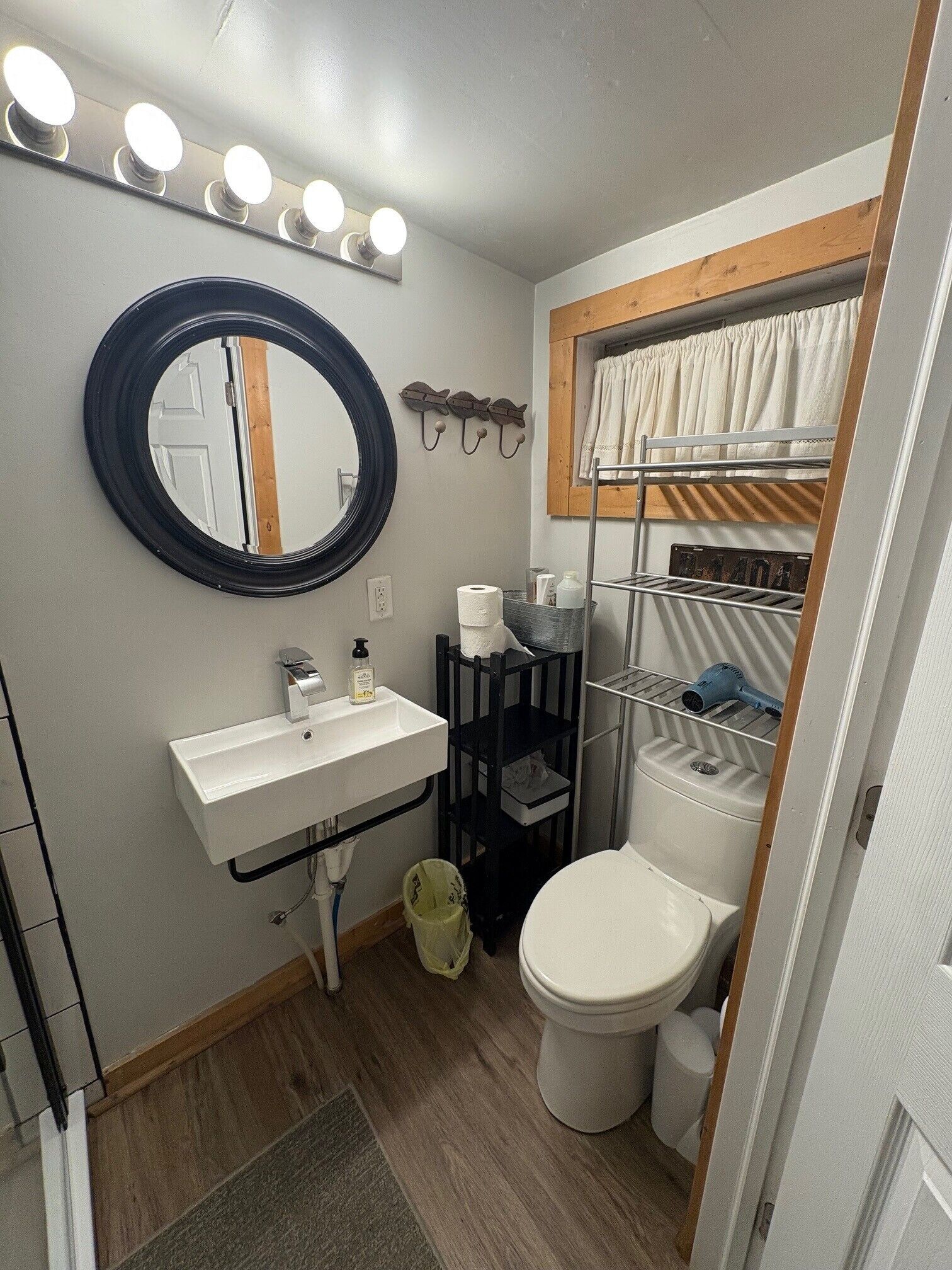 A compact bathroom with a round mirror, vanity, toilet, and a small shelf with toiletries.