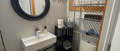 A compact bathroom with a round mirror, vanity, toilet, and a small shelf with toiletries.