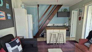 Ein Wohnzimmer mit einer schwarzen Couch, einem weißen Couchtisch und einem roten Sessel. Im Hintergrund ist eine Küche mit einer Holztreppe zu sehen.