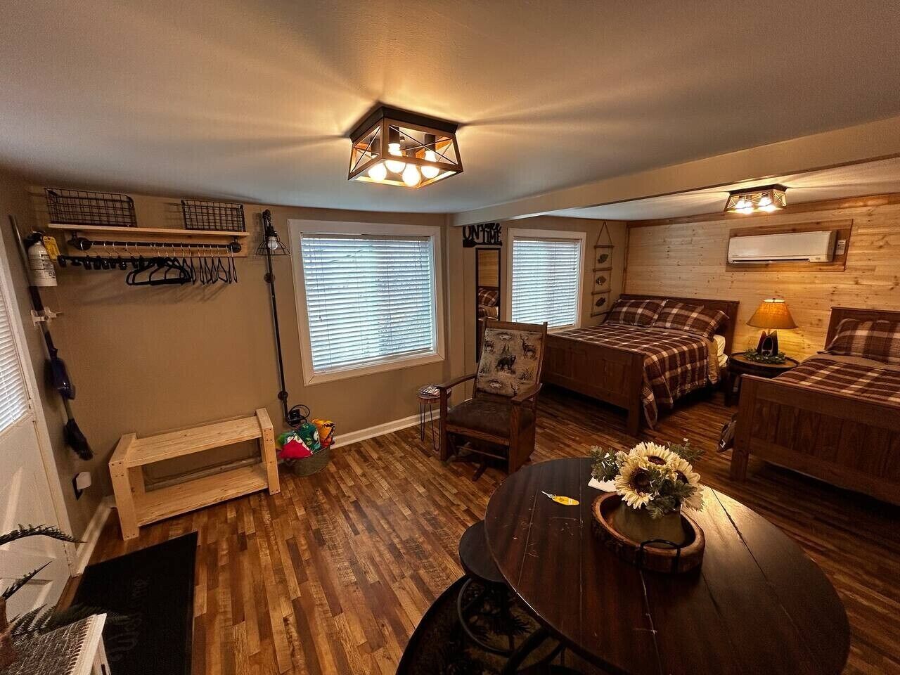 A cozy living room with a wooden floor, a round table with a vase of sunflowers, and a bed with a plaid bedspread.