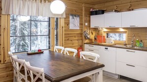 Interior de una cabaña de madera con una mesa de comedor, sillas blancas, una cocina con armarios blancos y una ventana con vistas nevadas.