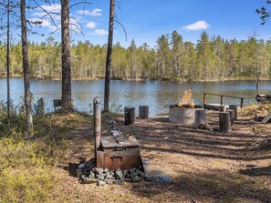 A campsite with a fire pit, a grill, and a concrete fire ring by a lake surrounded by trees.