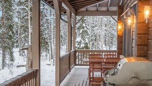 Terraza de madera con una parrilla y asientos exteriores, con vista a un bosque nevado.