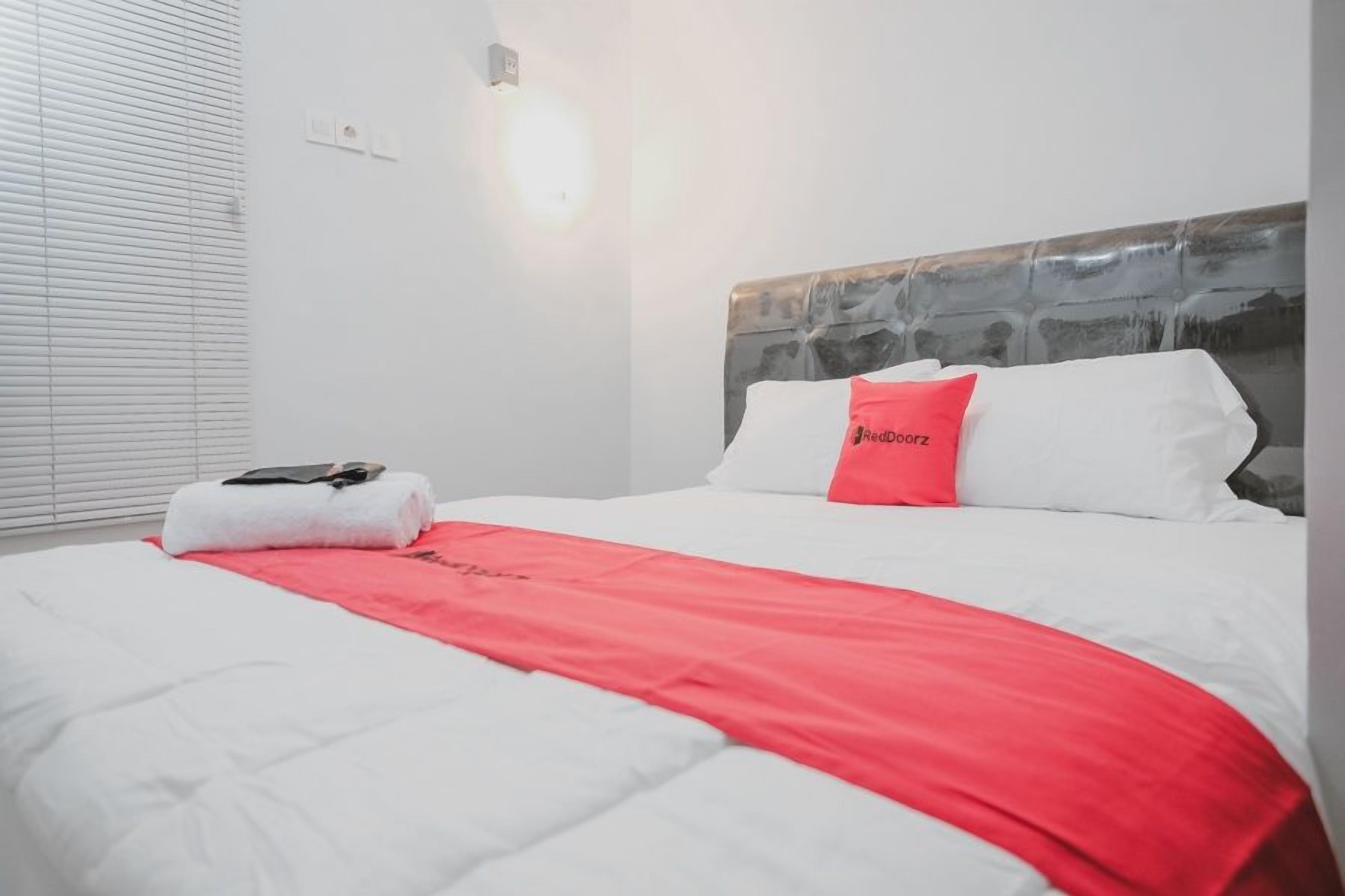 Habitación de hotel con cama, una manta roja, una almohada blanca y una toalla blanca sobre la cama.