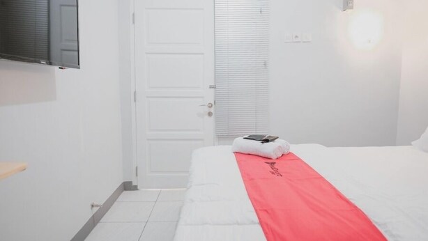 Quarto de hotel com cama, porta branca, janela com persianas, televisão montada na parede e mesa de cabeceira.