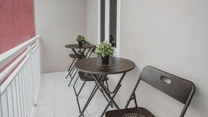 Uma varanda com guarda-corrimão branco, parede vermelha e chão branco. Há duas mesas redondas com cadeiras e plantas em vasos.