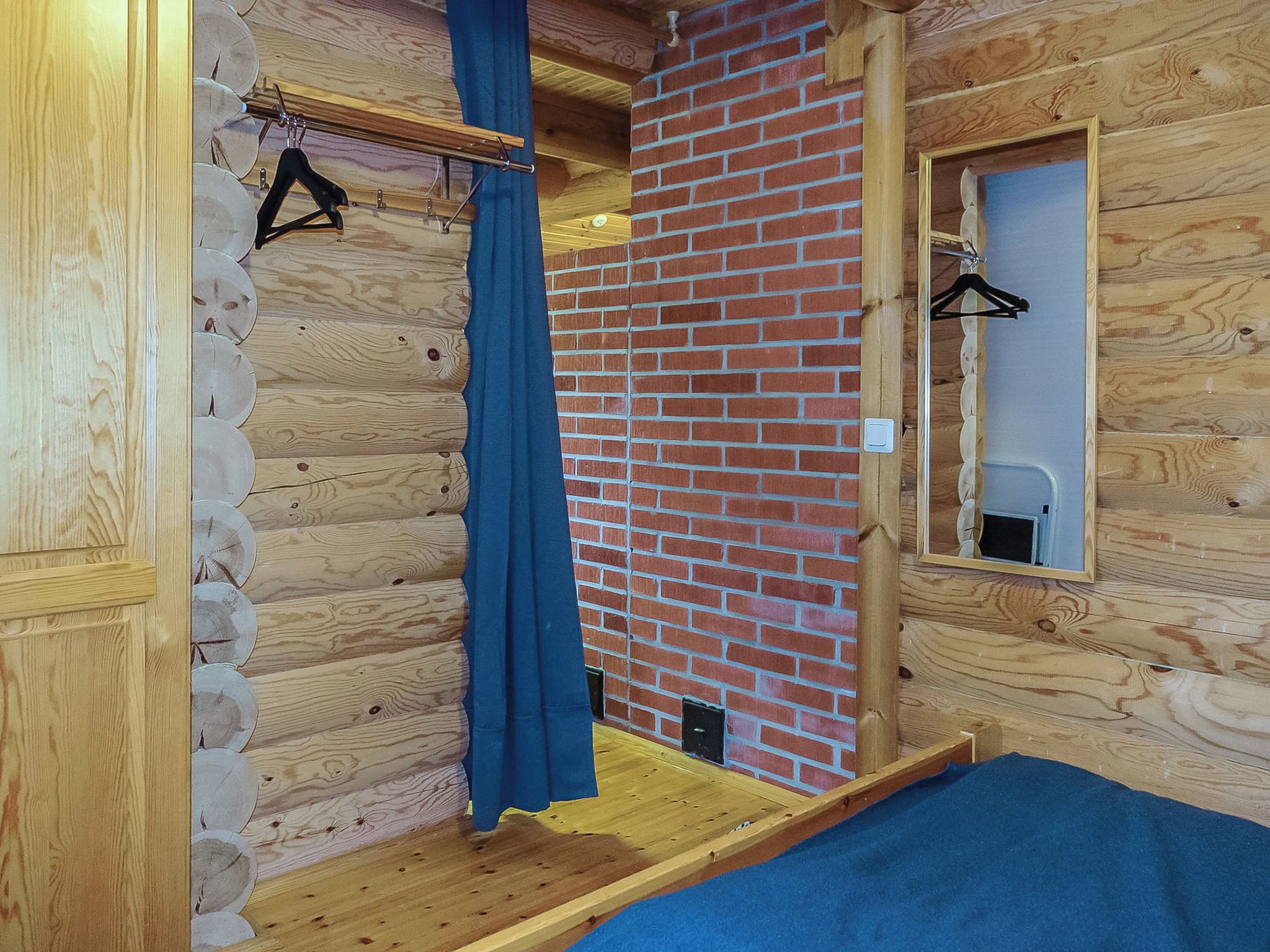 A wooden cabin interior with a brick wall, a wooden floor, a blue curtain, and a mirror on the wall.