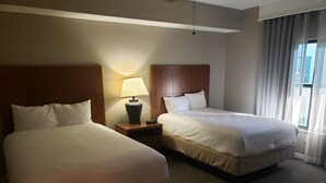 A hotel room with two single beds, a nightstand with a lamp, and a view of the city through large windows.