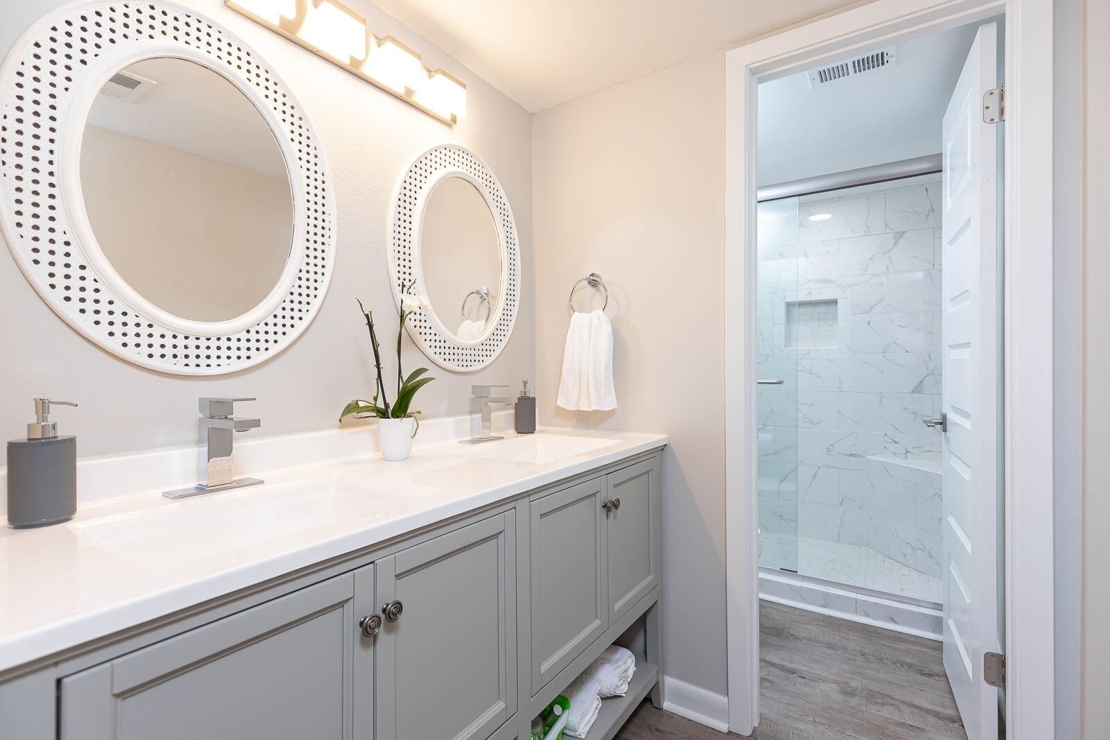 Une salle de bain moderne avec deux grands miroirs ronds, un meuble-lavabo blanc, une douche avec des portes en verre et une serviette accrochée au mur.