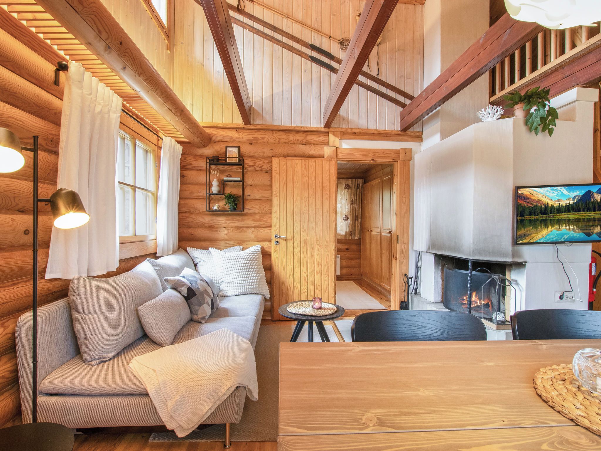 Un interior de cabaña acogedor, con techo de madera, un sofá con cojines, una pequeña mesa redonda y una chimenea.