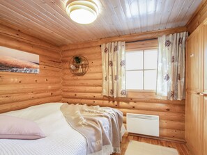 A log cabin bedroom with a bed, a window with curtains, a radiator, and a wall-mounted shelf with a plant.