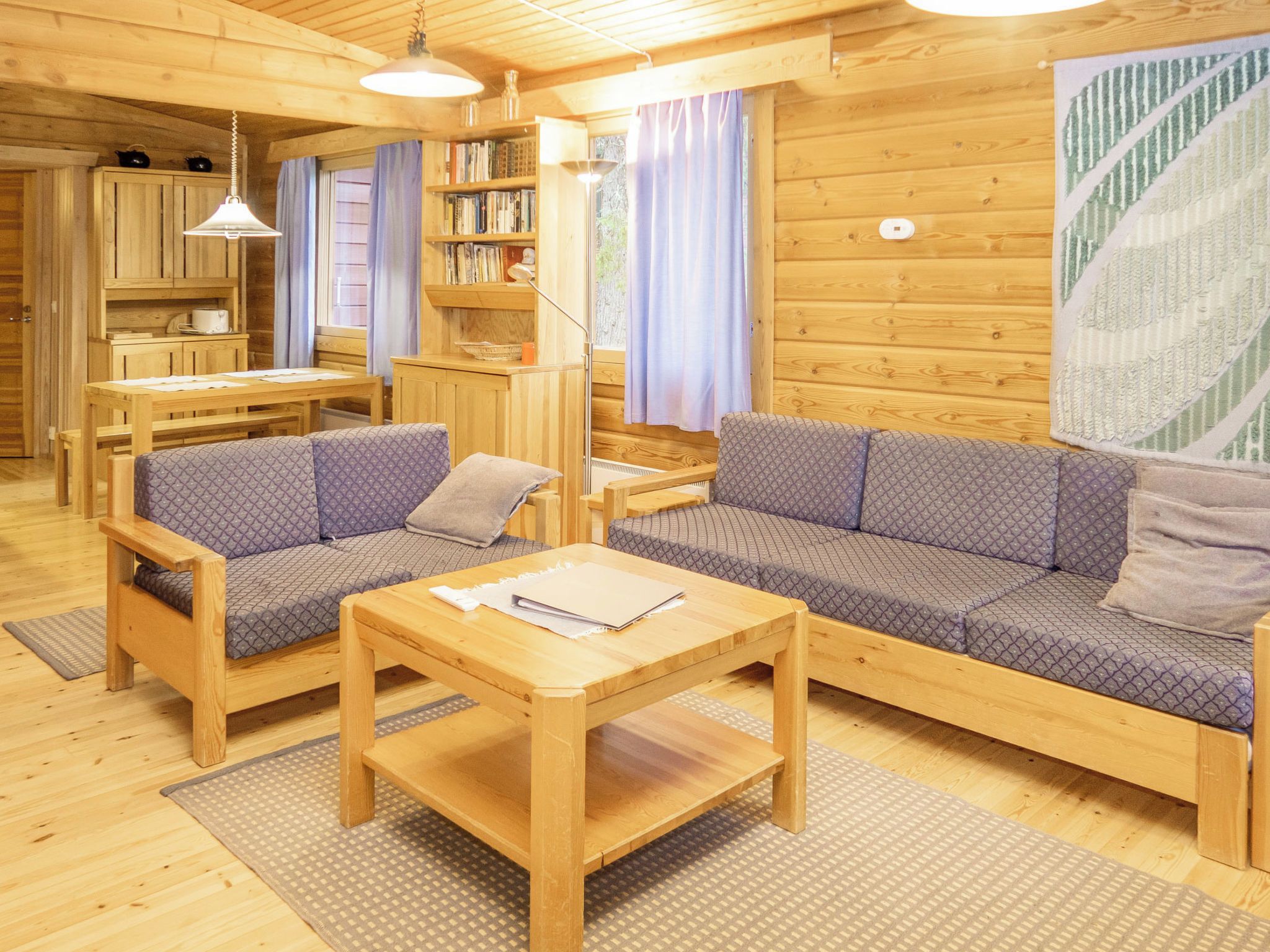A cozy cabin interior with a wooden dining table, two sofas, a bookshelf, and a window with curtains.
