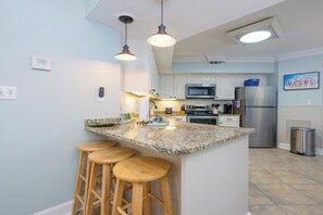 A kitchen with a granite countertop, wooden stools, a stainless steel refrigerator, and a microwave. - Three Bedroom ~ Gulf Front ~ Pelican Pointe! (Orange Beach)