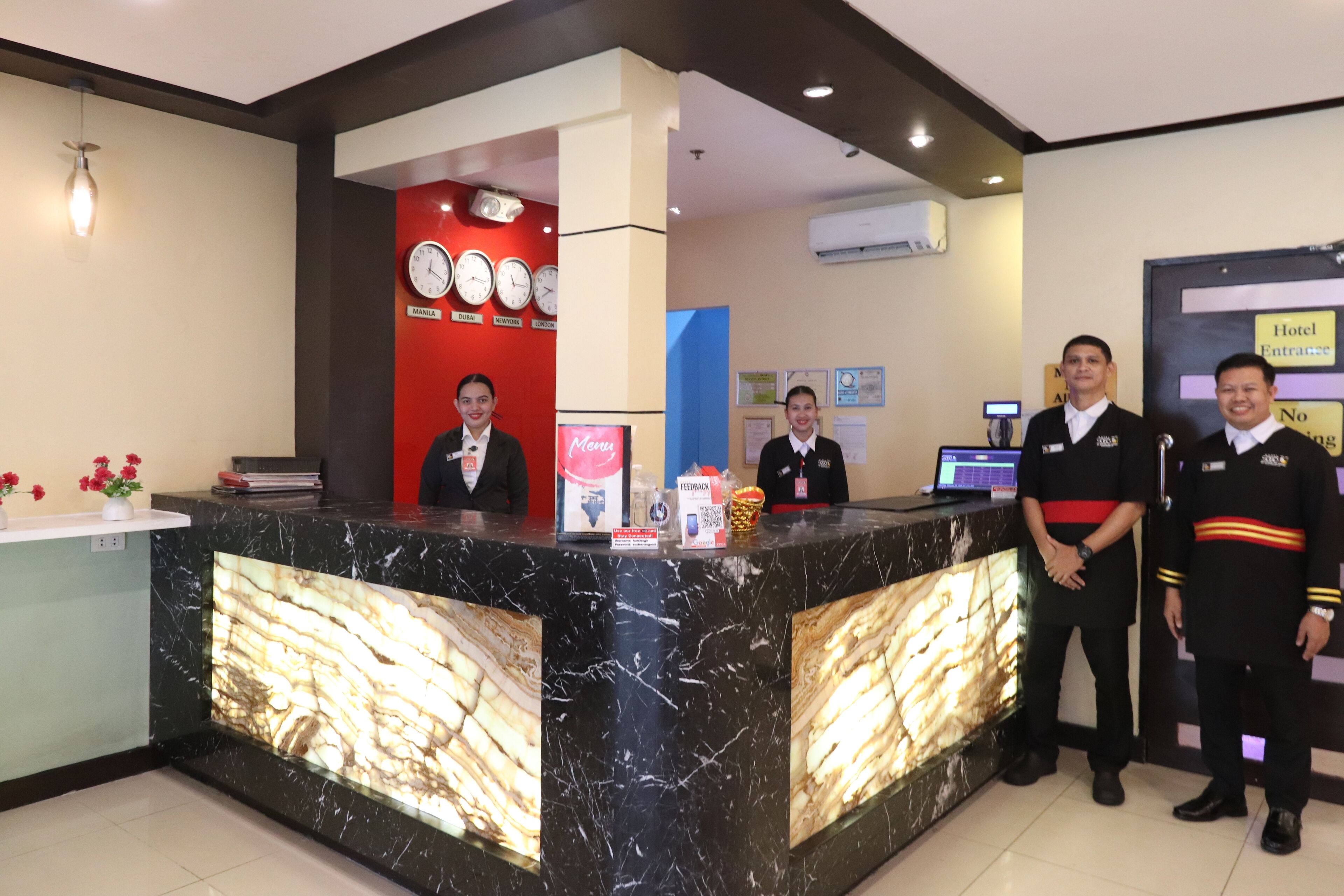 A hotel reception desk with a marble counter, clocks displaying time in various cities, and a sign indicating the hotel entrance.