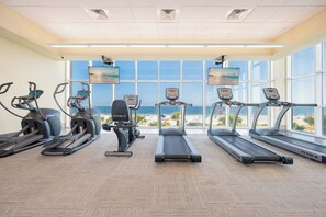 Academia com esteiras, bicicletas ergométricas e máquina de remo, com vista para uma praia.