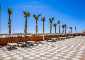 A beachside terrace with palm trees, sun loungers, and a tiled floor.
