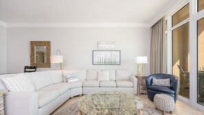 Una sala moderna con un sofá seccional blanco, una mesa de centro de vidrio con base de roca, un sillón de cuero azul y un amplio ventanal con cortinas.