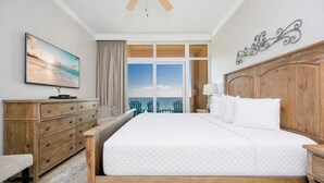 Habitación de hotel con una cama grande, cabecero de madera, televisor en una cómoda, vista al océano y un balcón con sillas.