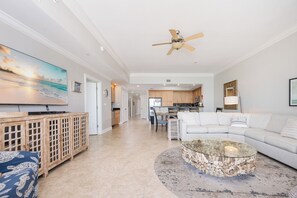 A spacious living room with a large white couch, a glass coffee table, and a wooden cabinet. A ceiling fan is visible, and the room leads to a kitchen area.