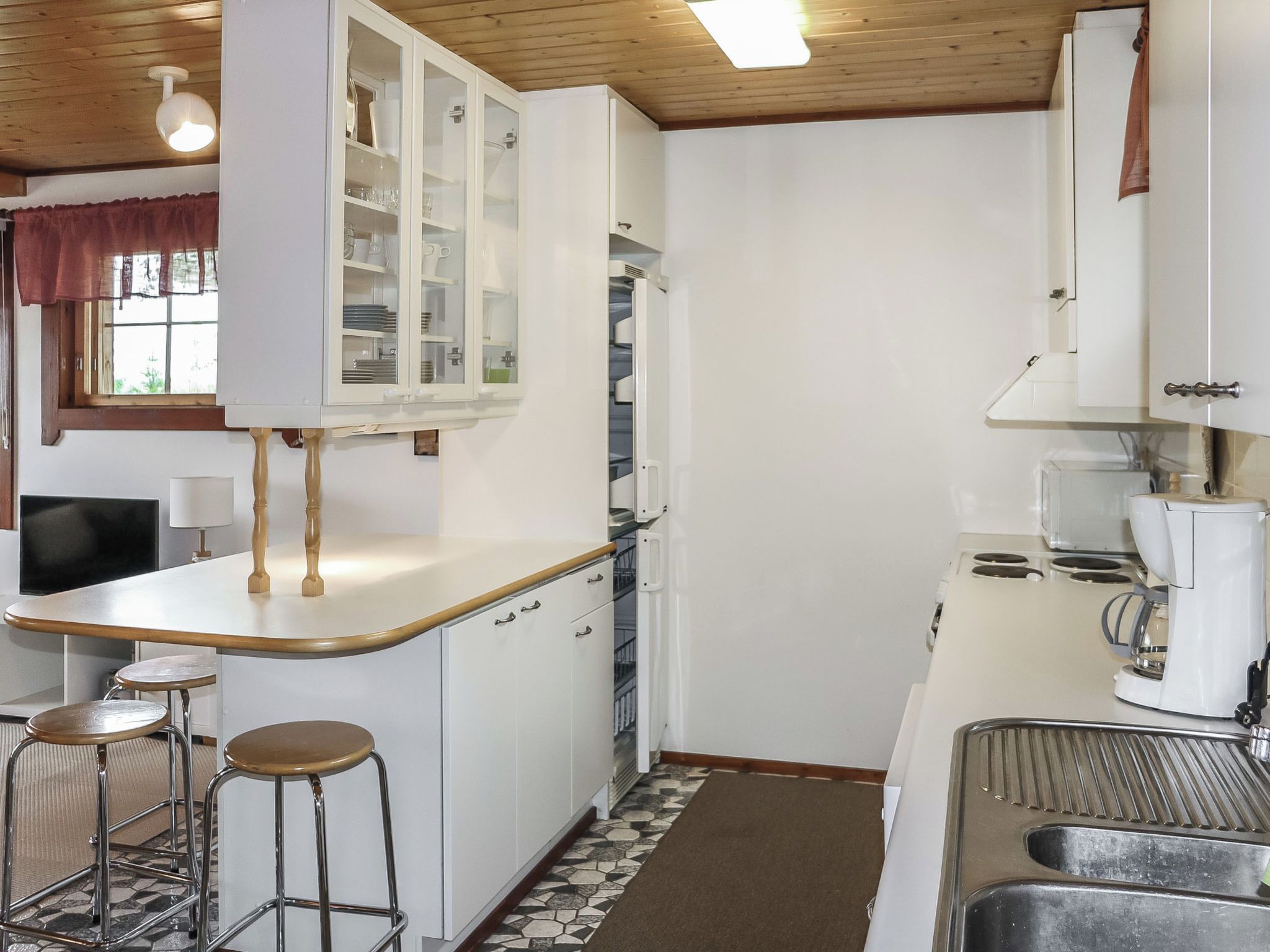 Cocina funcional con gabinetes blancos, estufa integrada y cafetera. Hay dos taburetes y una ventana con cortinas.