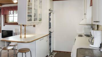 Cocina funcional con gabinetes blancos, estufa integrada y cafetera. Hay dos taburetes y una ventana con cortinas.