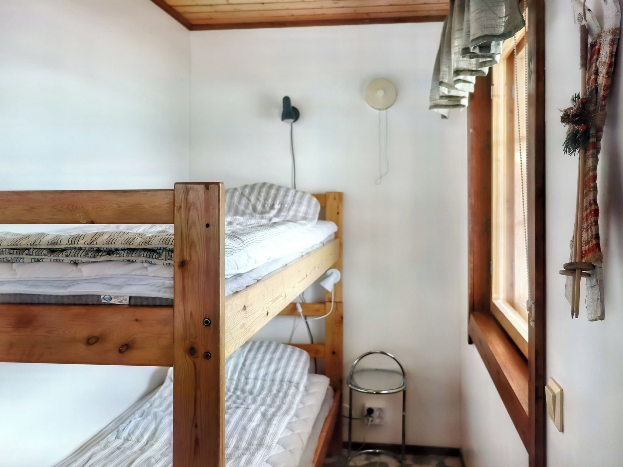 A small, compact room with a bunk bed, a bedside table, a wall-mounted lamp, and a window with curtains.
