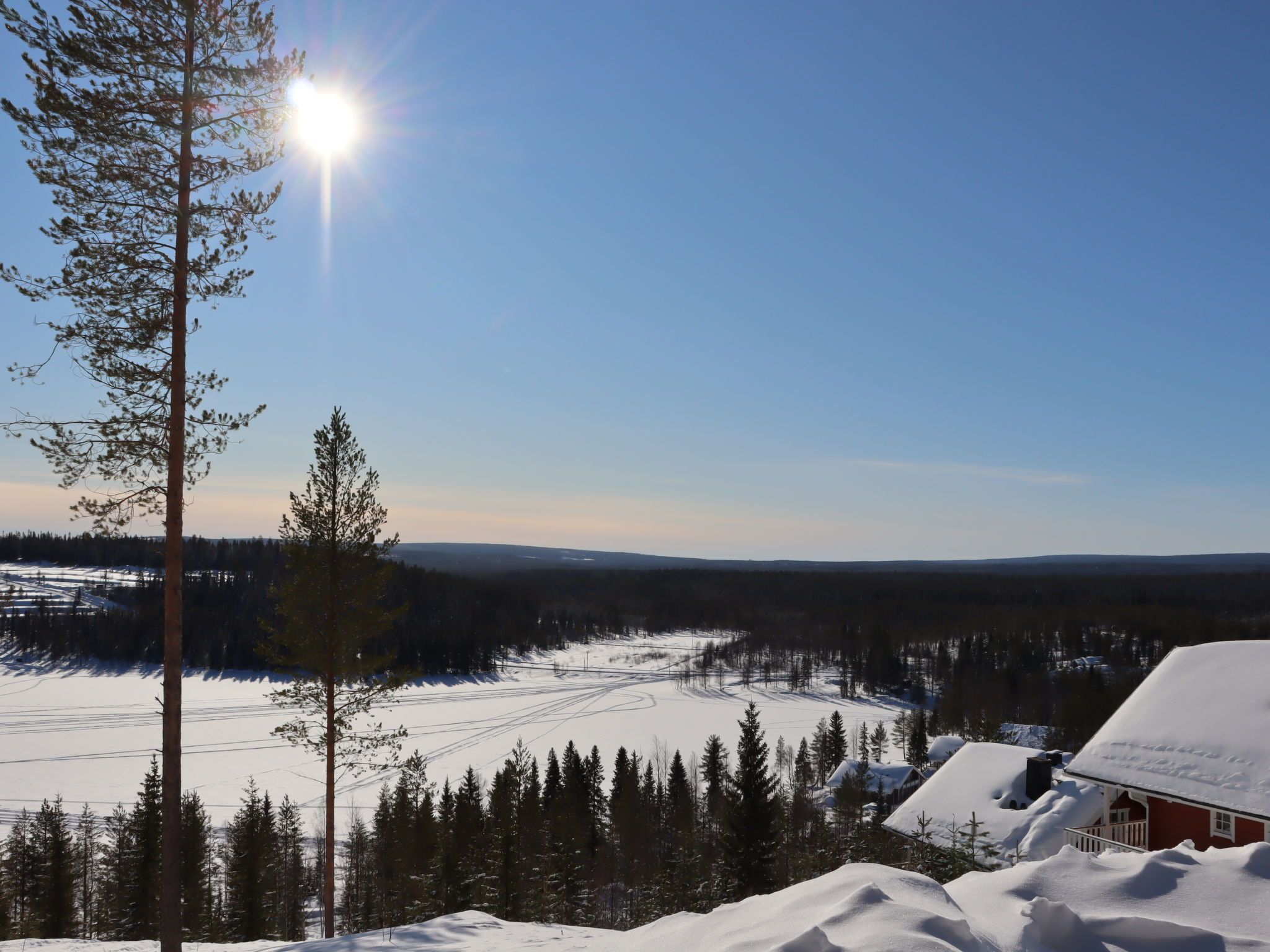 Un paysage enneigé, avec un ciel dégagé, quelques arbres et une maison dont le toit est recouvert de neige.