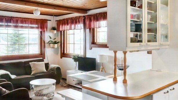 A cozy living room with a sofa, a coffee table, and a TV. There's a kitchen area with cabinets and a countertop.