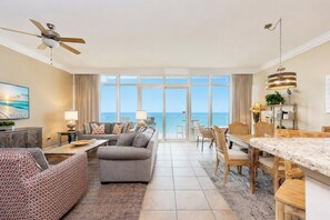 A spacious living room with a large window, a ceiling fan, and a dining area with a view of the ocean.