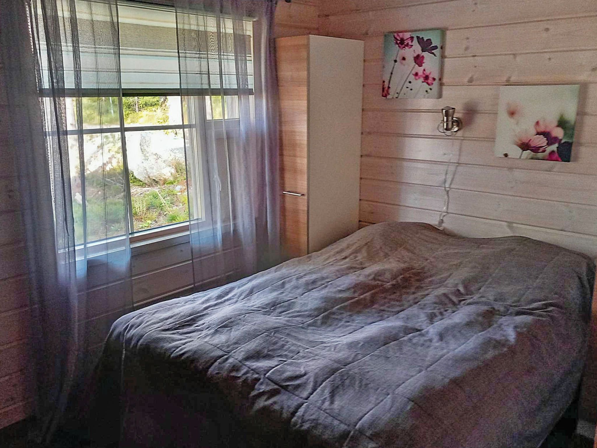 A bedroom with a bed, a window with curtains, a wardrobe, and two wall-mounted paintings.