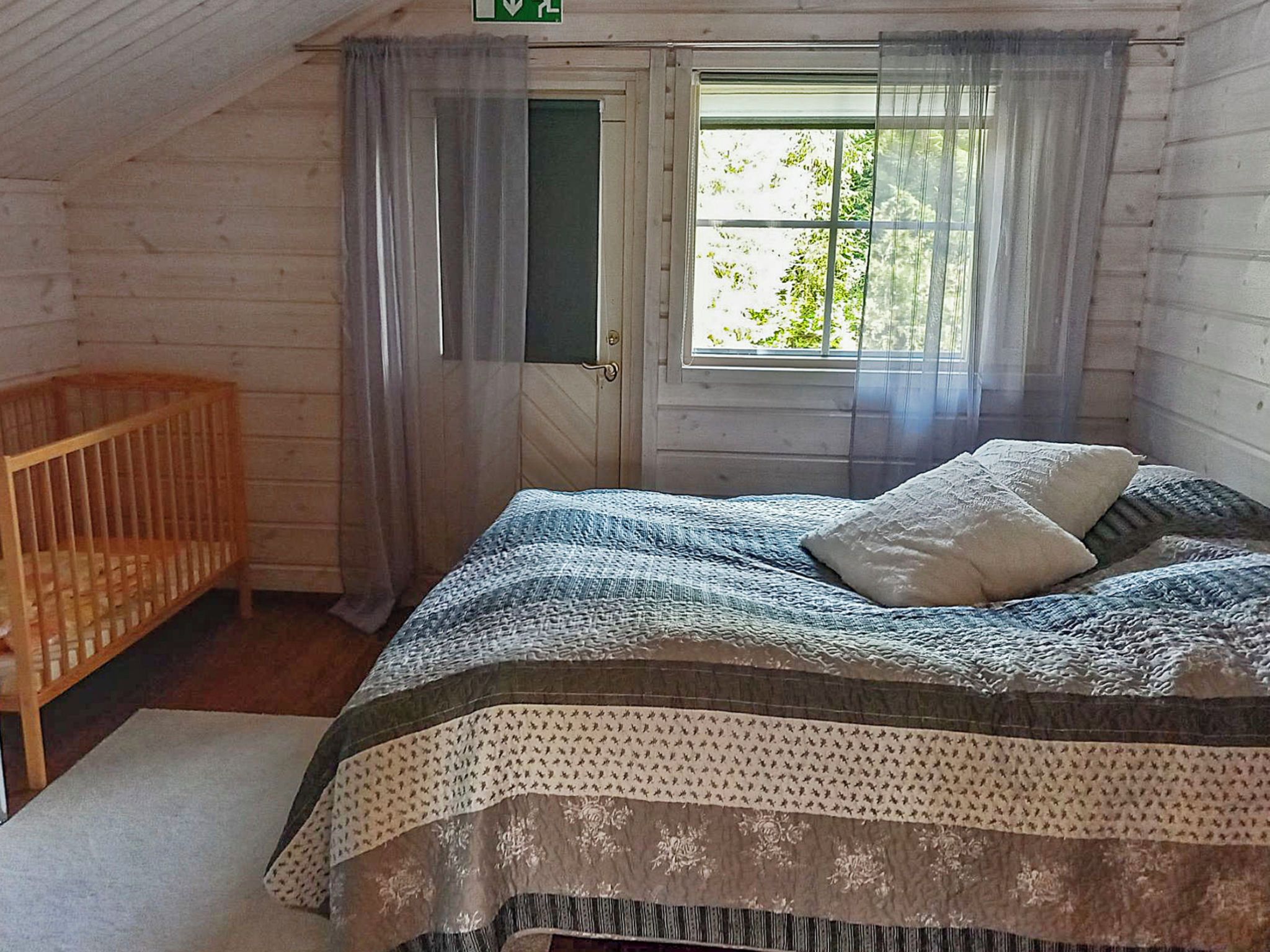 Une pièce avec un lit simple, un berceau en bois, une fenêtre avec des rideaux et une porte.