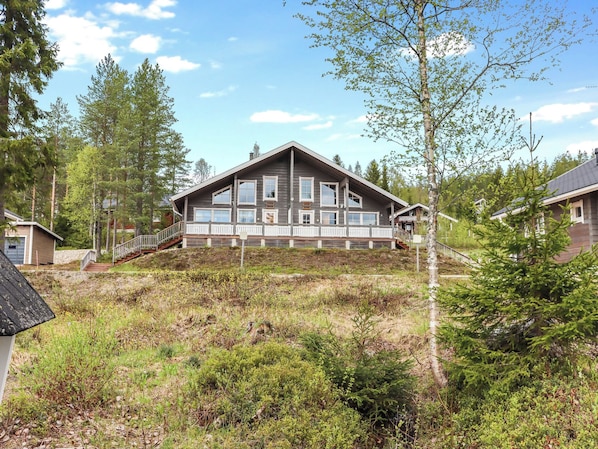 A large two-story log cabin with a balcony, surrounded by dense forest and a grassy area.