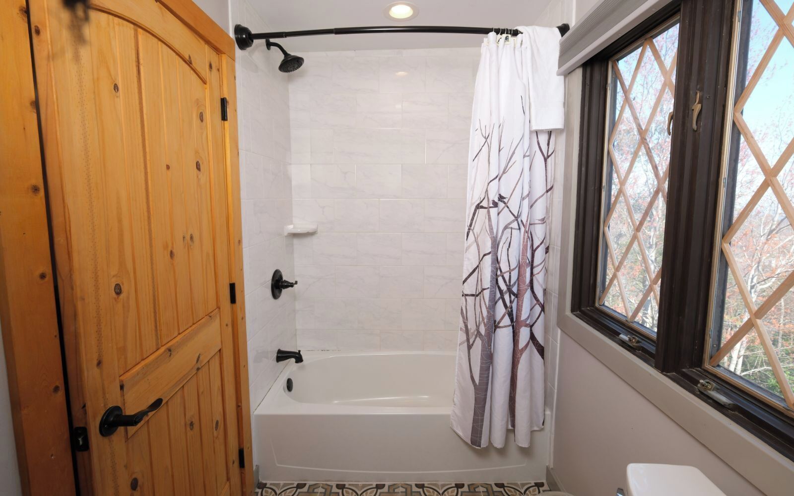 A bathroom with a wooden door, a bathtub, a shower curtain with a tree pattern, and a window with a view of trees.