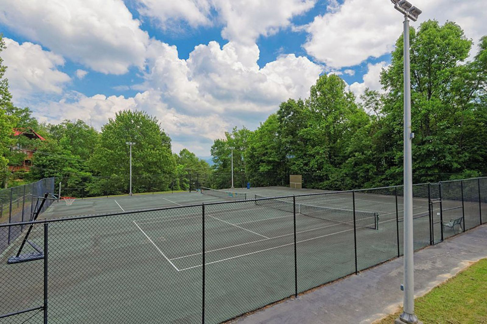 Una cancha de tenis al aire libre con una cerca de malla metálica, rodeada de árboles y un cielo despejado.