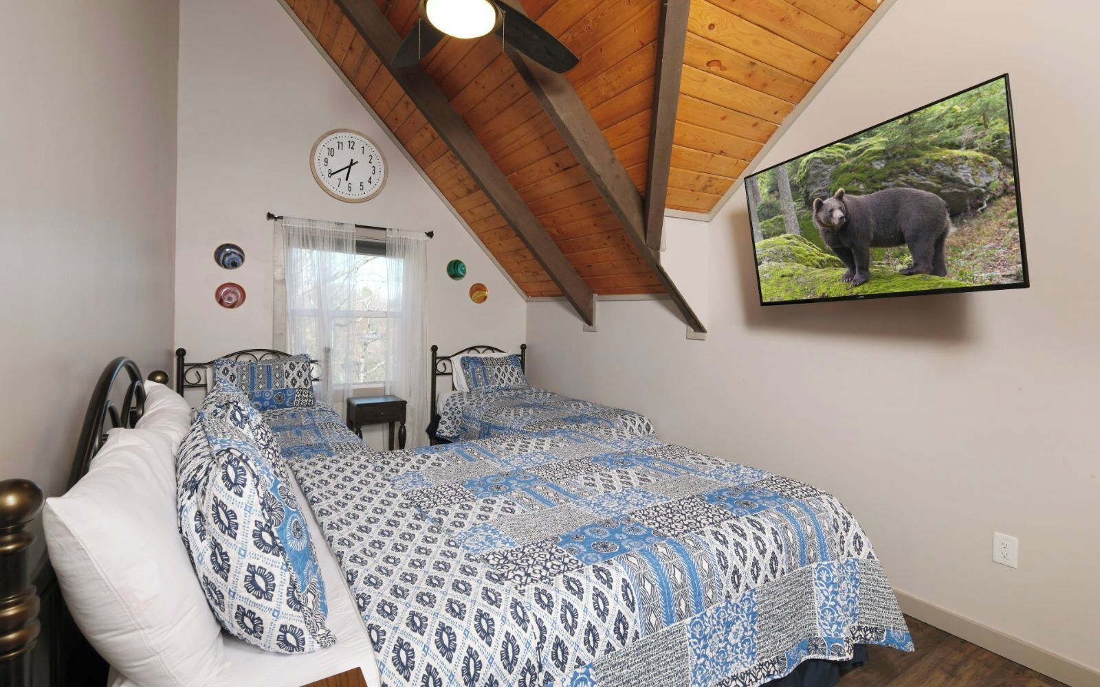 Quarto com teto inclinado, cama com roupa de cama estampada, relógio de parede, janela com cortinas e um quadro de urso na parede.
