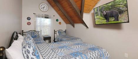 Quarto com teto inclinado, cama com roupa de cama estampada, relógio de parede, janela com cortinas e um quadro de urso na parede.