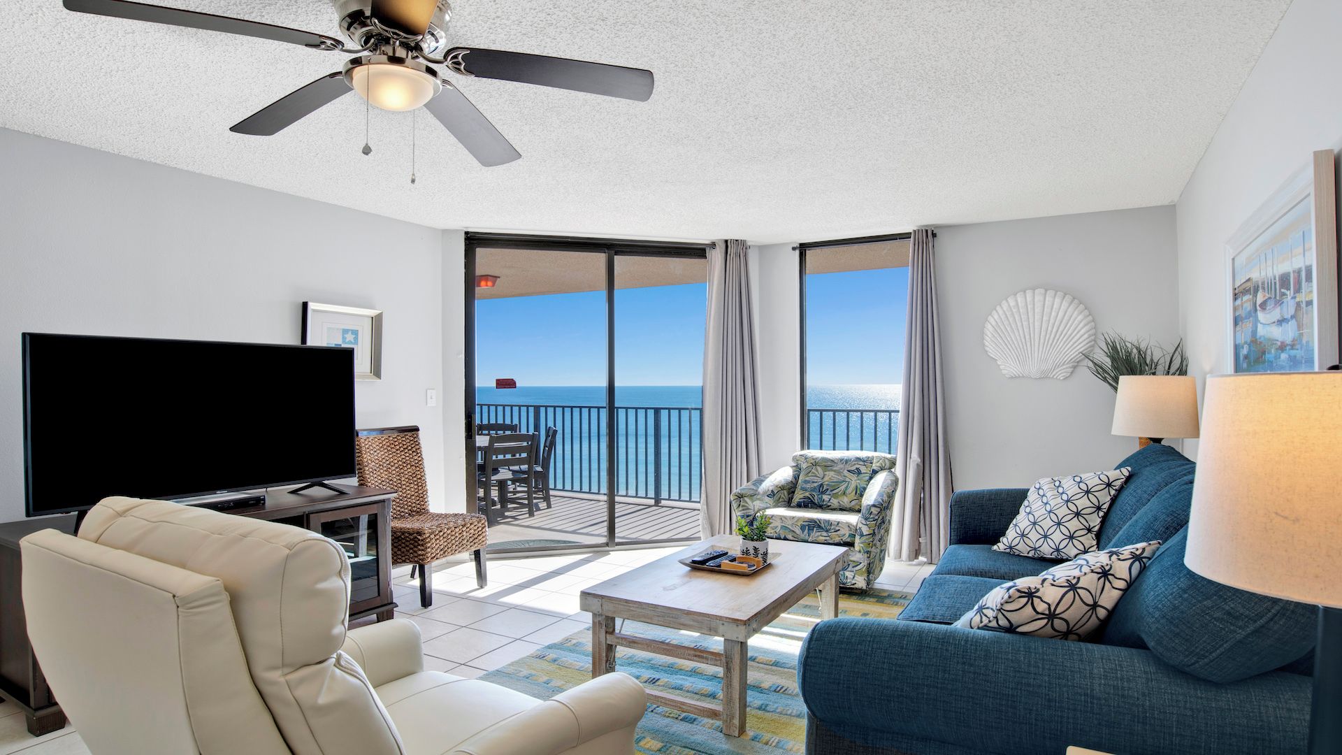 A living room with a TV, sofa, and coffee table. There is a balcony with a view of the ocean.