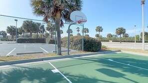An outdoor basketball court with a chain-link fence, palm trees, and a clear sky.