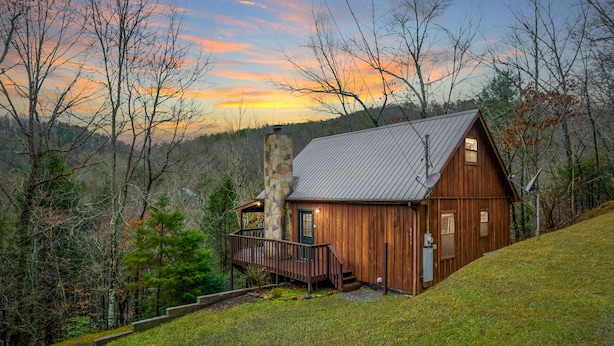 A wooden cabin with a stone chimney, surrounded by trees and a grassy hill. - River Ridge: Where Water Meets the Mountains (Pigeon Forge)