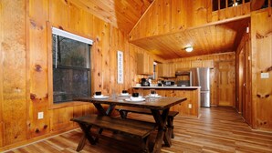 A wooden cabin interior with a wooden dining table set for four, a kitchen area with modern appliances, and a large window. - River Ridge: Where Water Meets the Mountains (Pigeon Forge)
