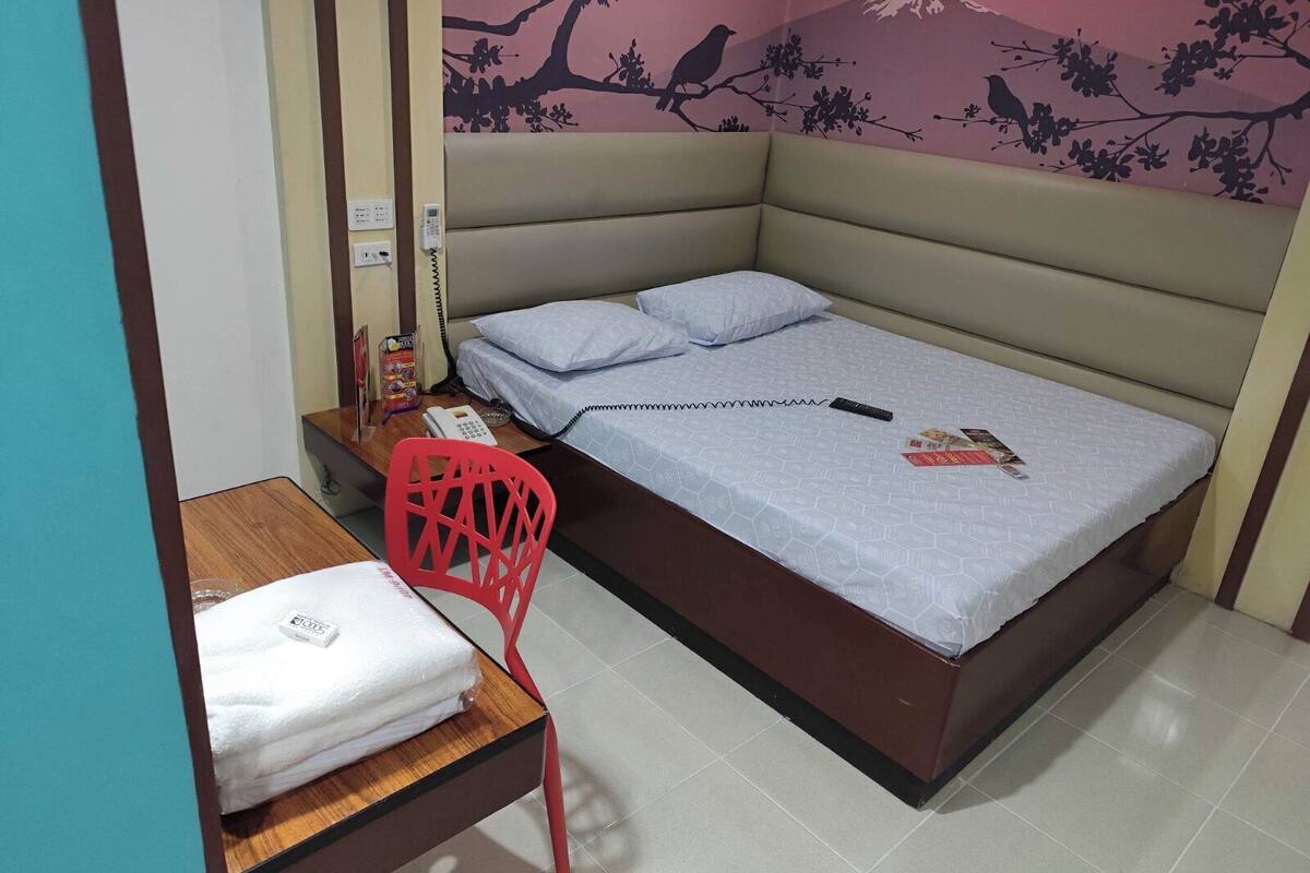 A small, neatly arranged bedroom with a bed, pillows, a red chair, a small table with a towel, and a wall with a bird and tree mural.